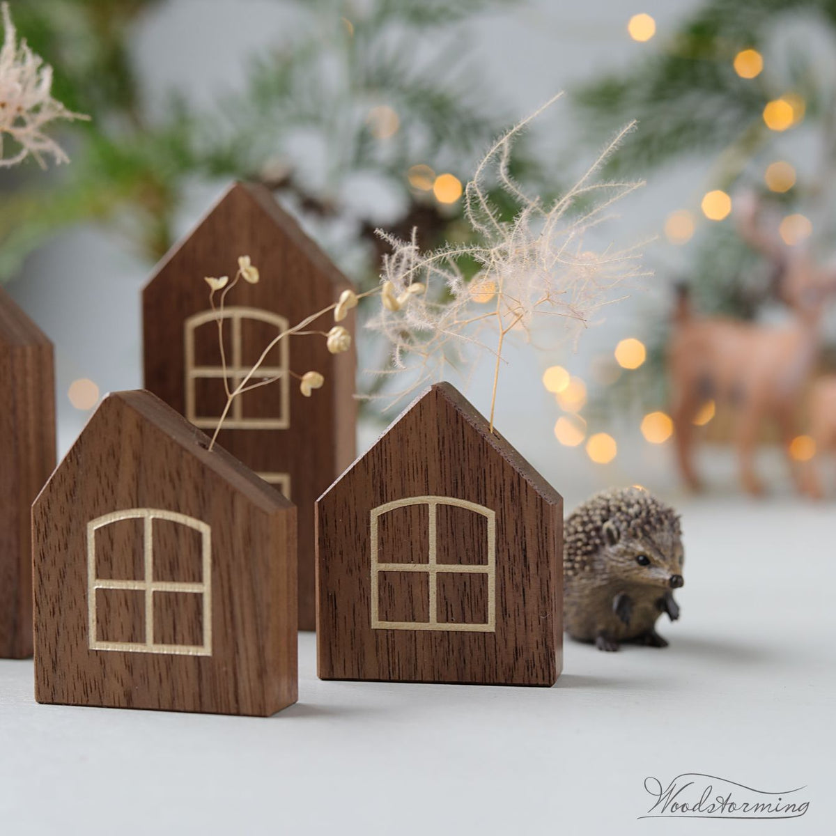 Three small wooden house-shaped ornaments with laser-cut window details, arranged on a light surface with decorative dried grass and a hedgehog figurine in a softly blurred holiday setting.