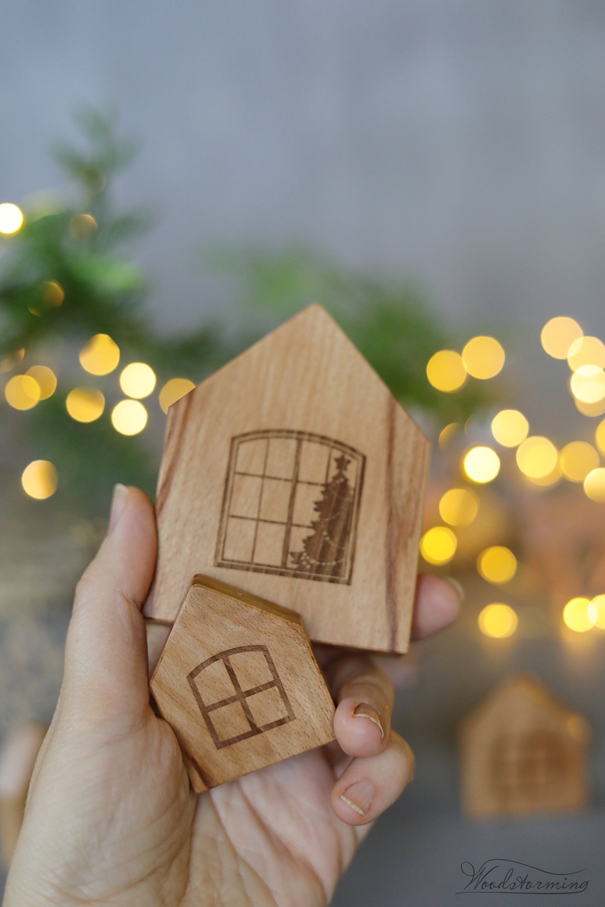 A hand holds two small wooden house-shaped items with engraved window and Christmas tree designs, set against a softly blurred background with warm bokeh lights and greenery.