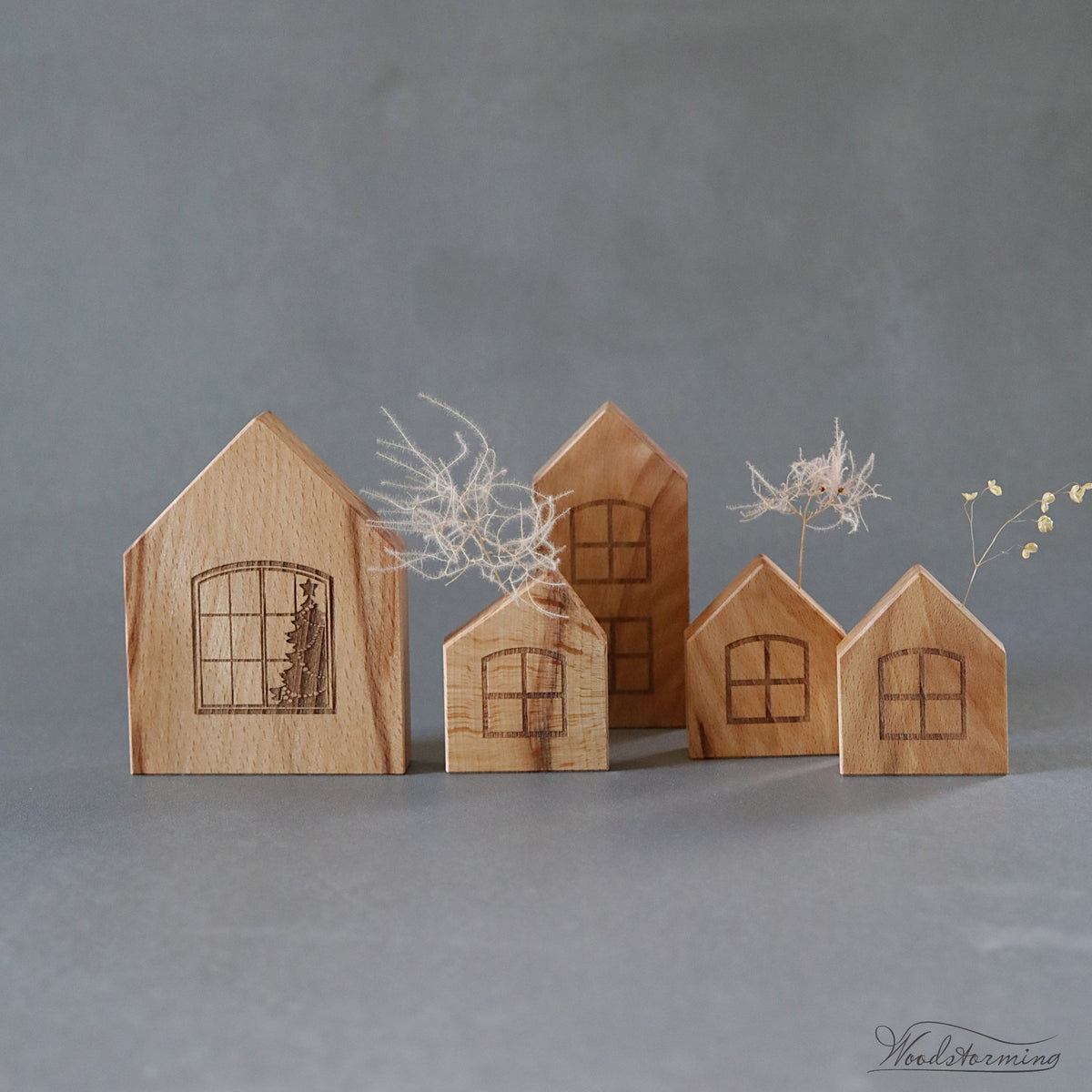 Five small wooden house-shaped ornaments with engraved window designs, arranged on a smooth gray surface against a gray background. Two small dried floral stems are placed behind and beside the houses.