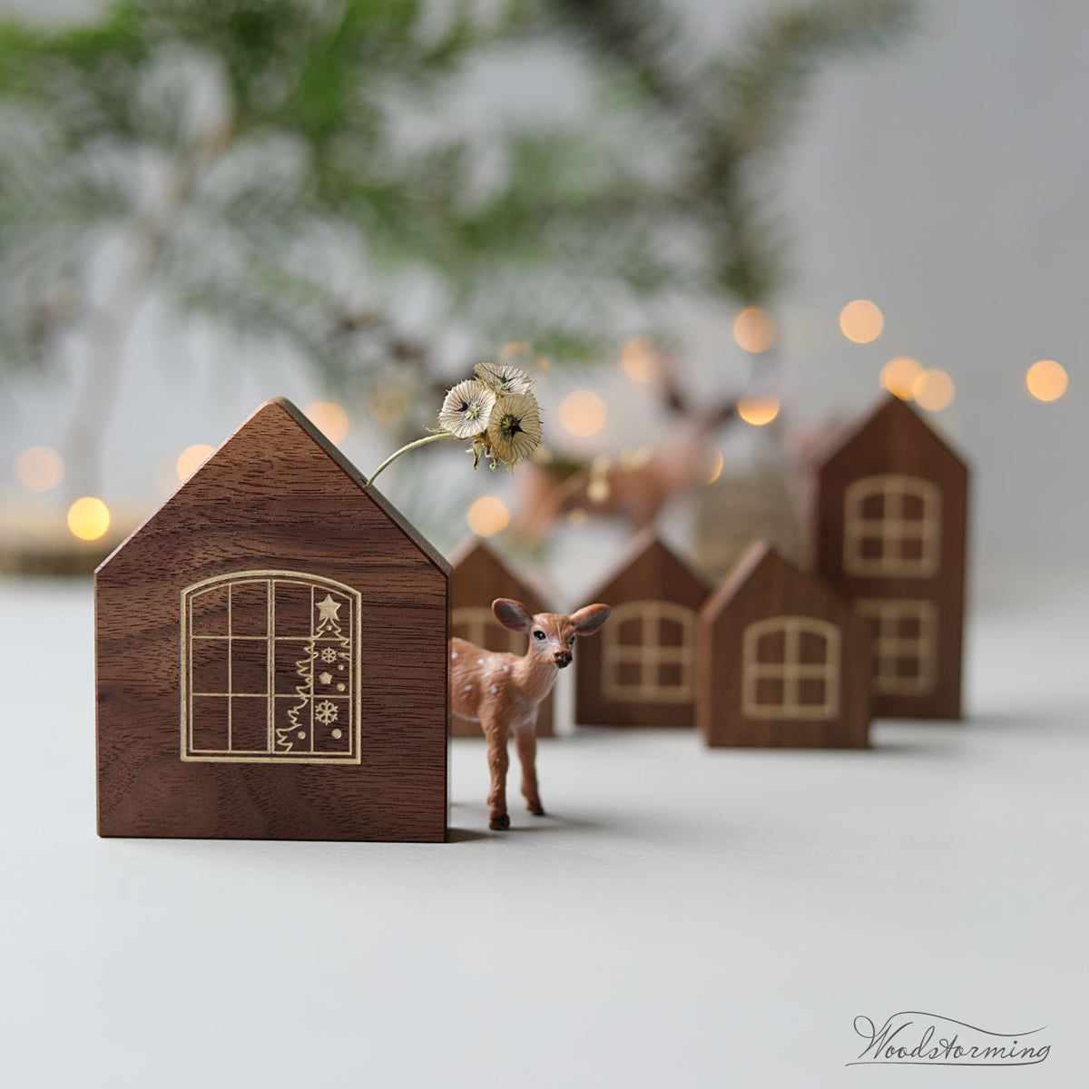 A set of handcrafted wooden house figurines with laser-engraved window details, displayed on a light surface with a small deer figurine and a dried flower stem in the foreground, set against a soft-focus background with warm bokeh lights.