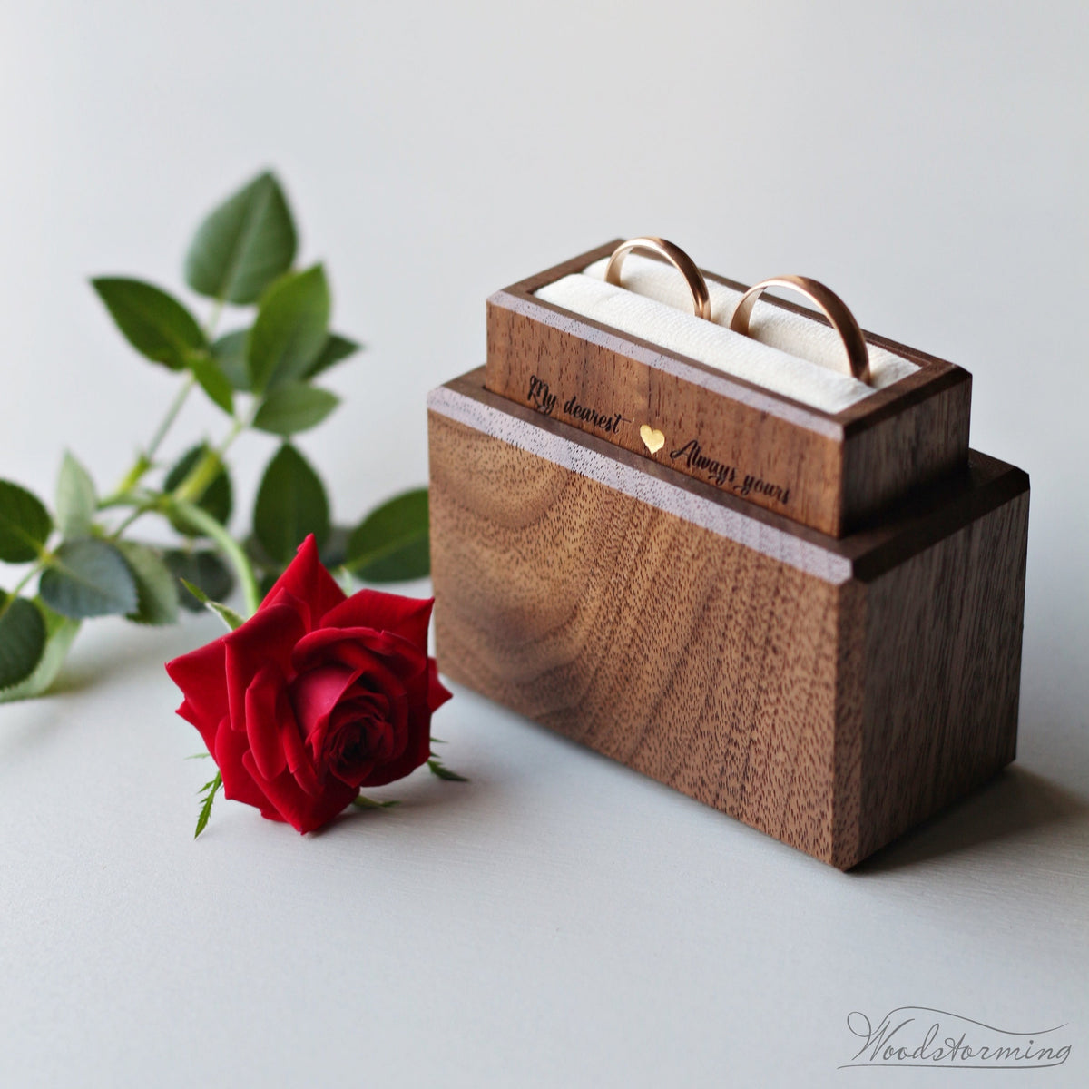 A minimalist wooden ring bearer box with elegant cream-white pillow, displaying two rings. The box is made of walnut, finished with ecological oil and wax, and has a personalized engraving.